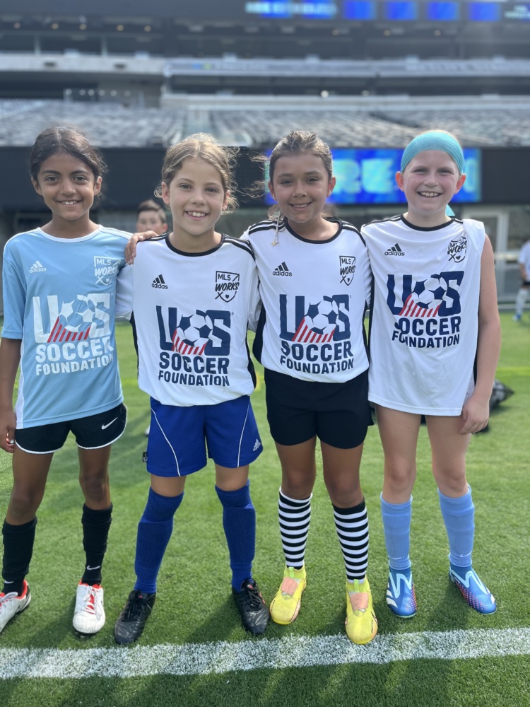 four young girls pose and smile for the camera with their arms around each other, each about ten years old. the girl on the left is wearing a sky blue U.S. Soccer Foundation jersey. the three girls to her left are each wearing a white jersey.